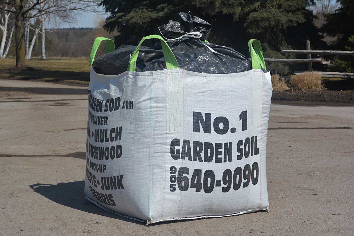 Large white bag of garden soil with green handles sits on pavement, surrounded by trees and a grassy area in the background.