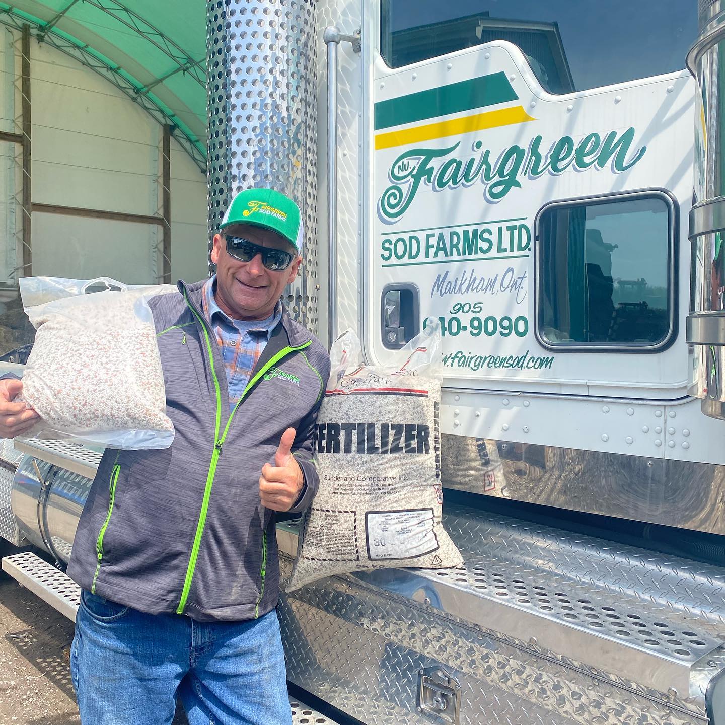 A person in a green cap and sunglasses stands beside a Fairgreen Sod Farms truck, holding bags of fertilizer, smiling and giving a thumbs-up.