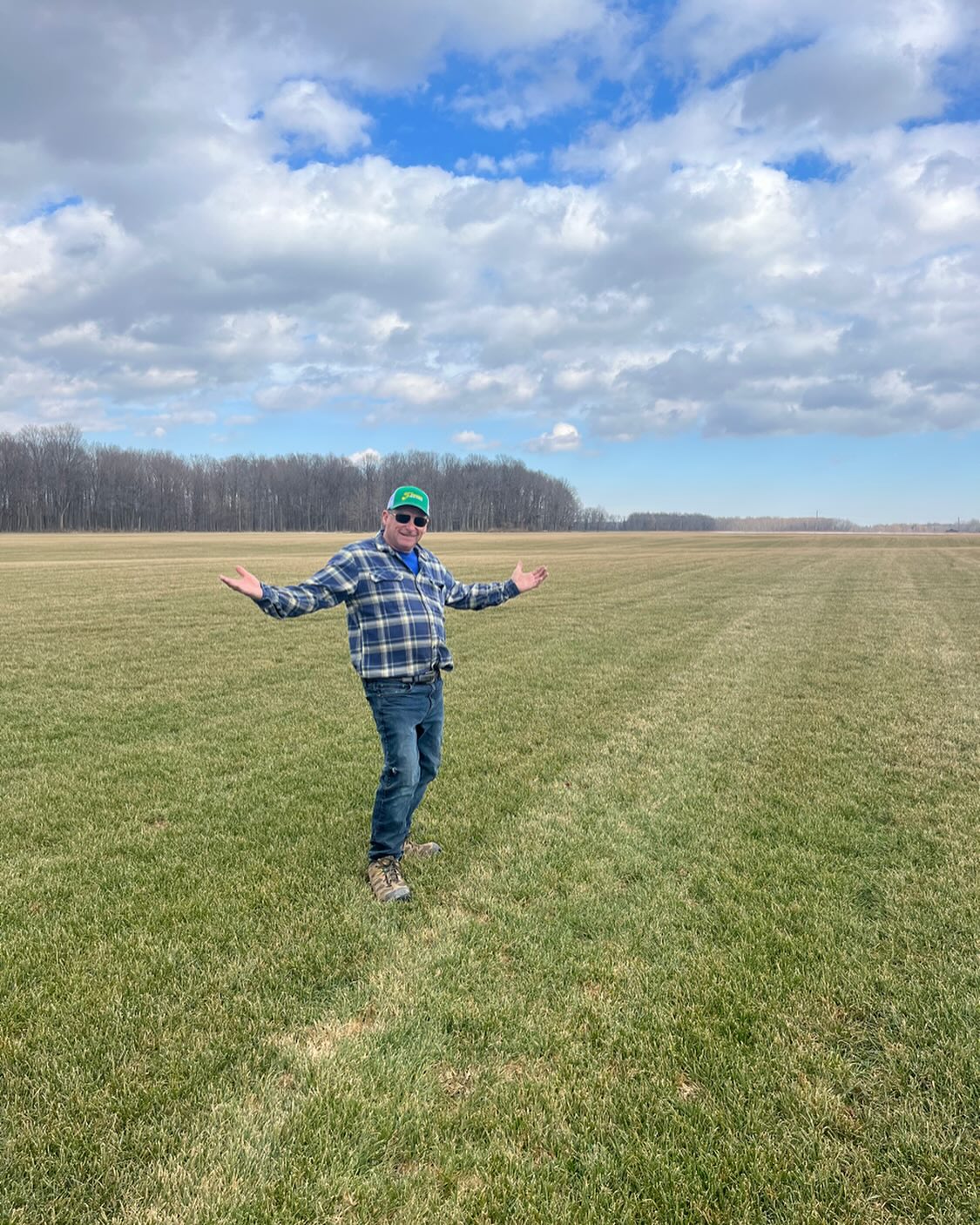 A person in a green cap stands in a vast grassy field, with trees and a cloudy blue sky in the background.