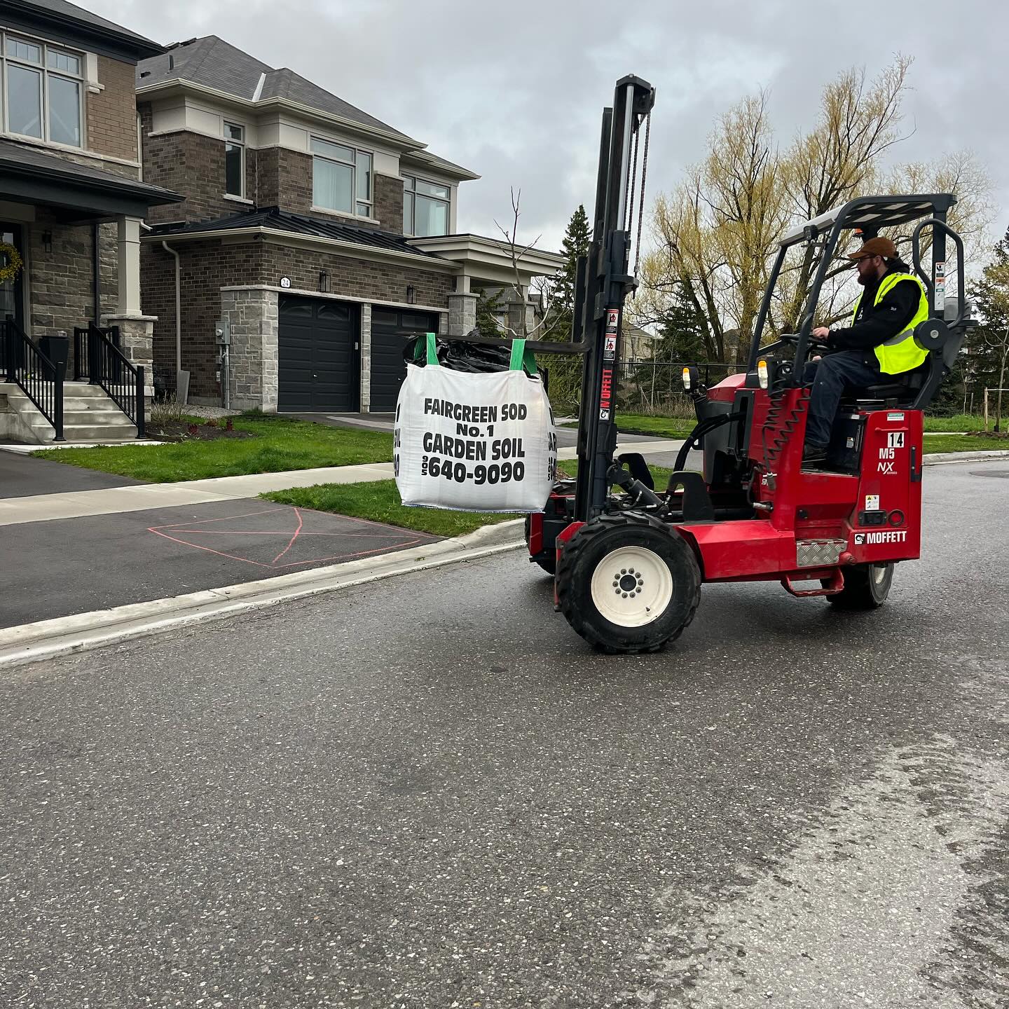 A person operates a forklift carrying garden soil on a suburban street, with modern houses visible in the background.