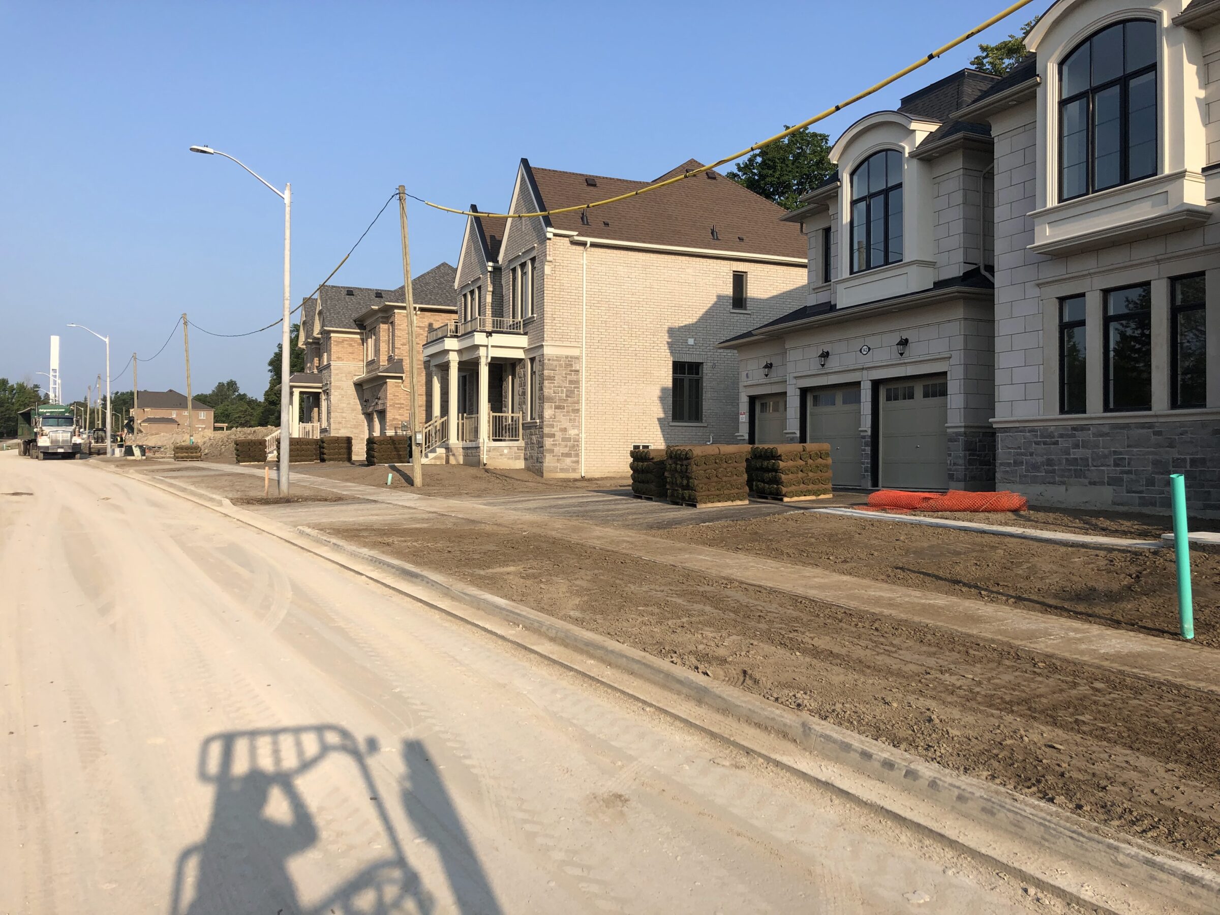 A row of newly constructed suburban homes under clear skies, with grass sods stacked outside. A street cleaner truck appears in the distance.