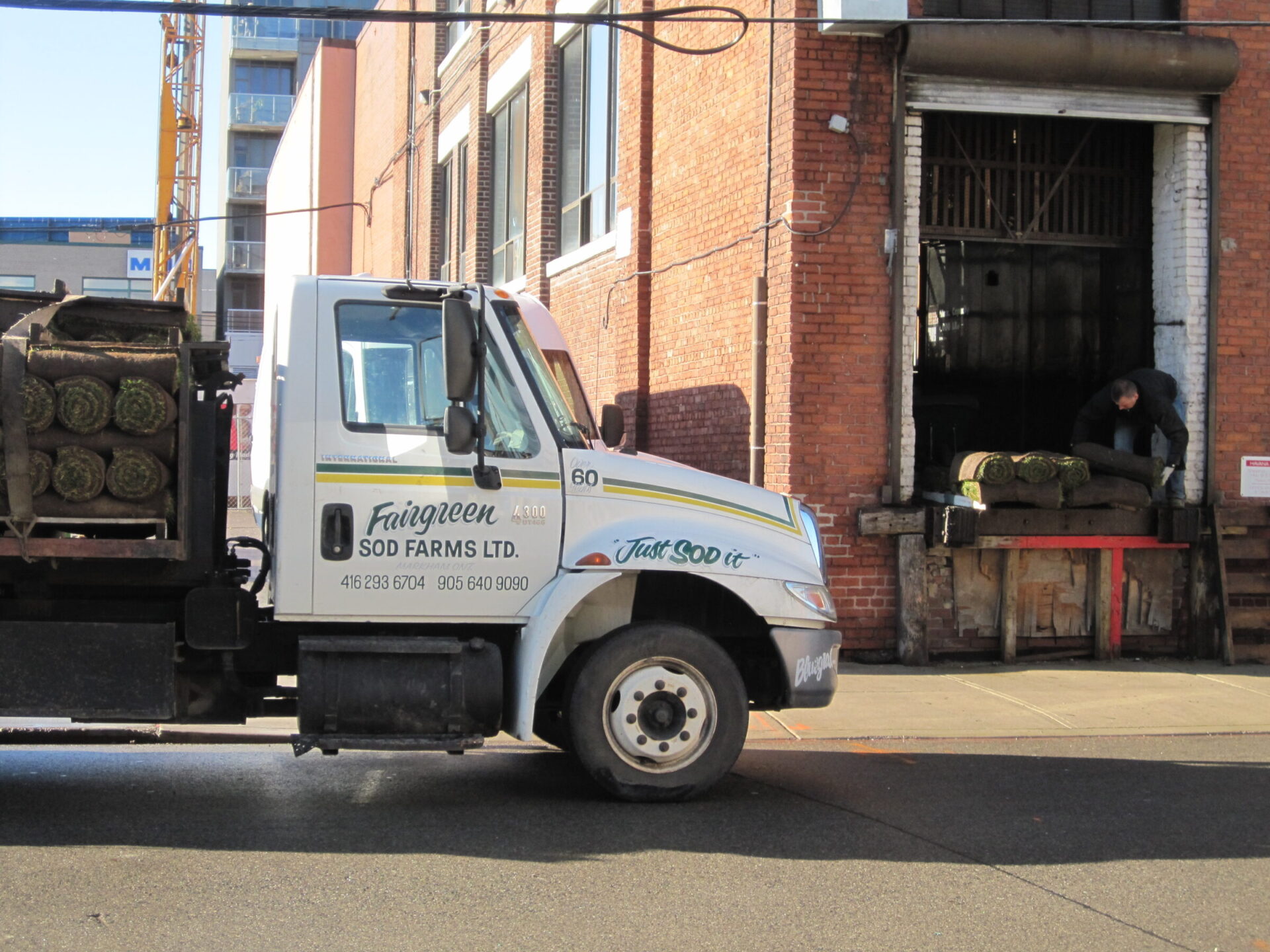 A truck from Fairgreen Sod Farms, loaded with sod, parked outside a red brick warehouse. A person loads sod onto a platform.