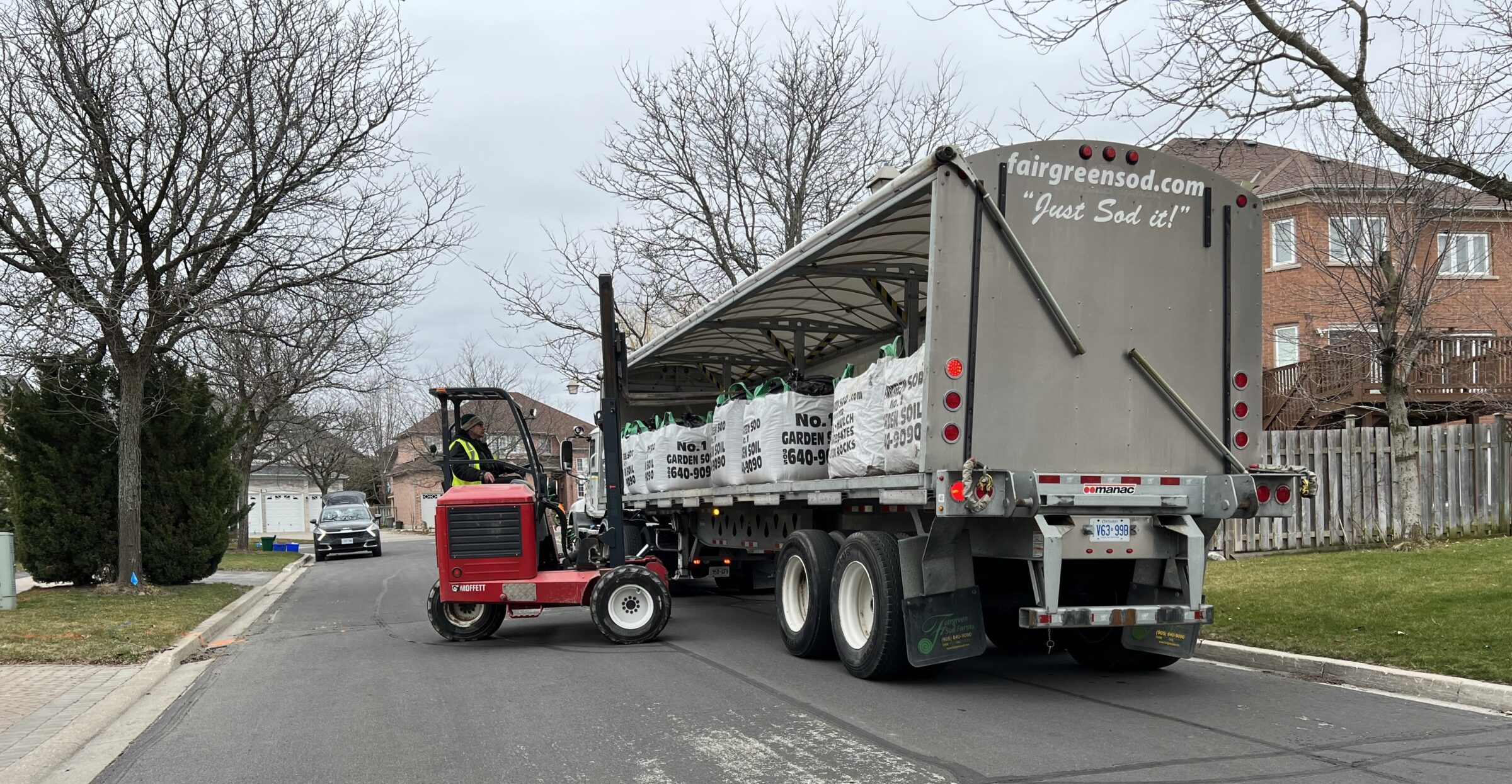 A truck loaded with garden soil bags is parked on a suburban street. A person operates a small forklift nearby.