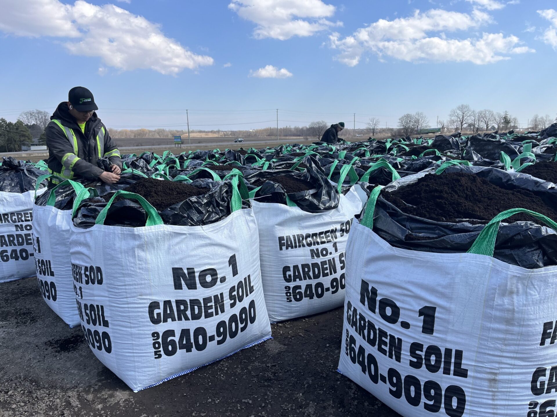 A person inspects large bags of garden soil outdoors. Overcast sky, trees, and fields in the background. No recognizable landmarks visible.