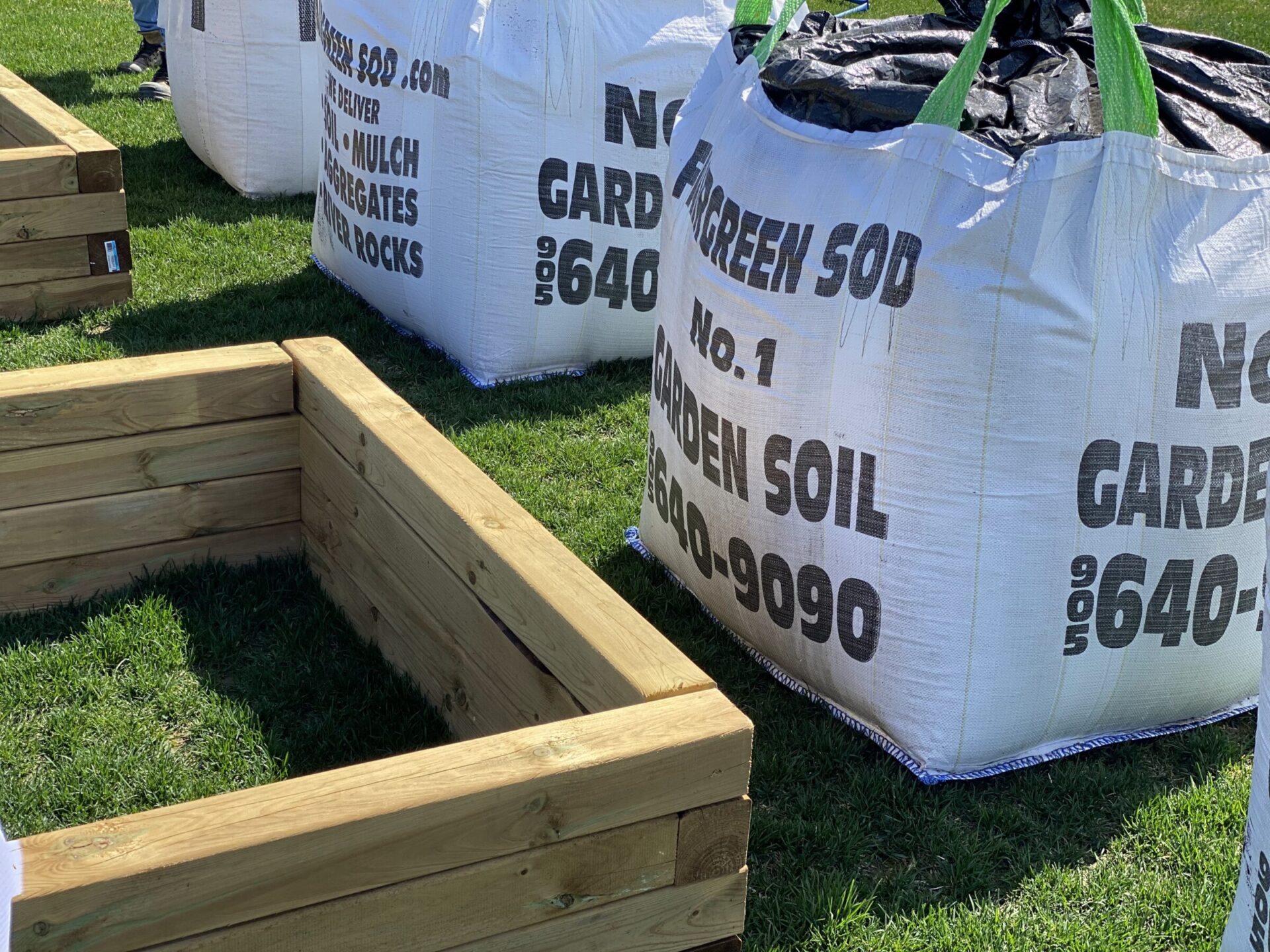 Wooden garden beds and large soil bags on grass. Two persons partially visible near the bags. Bright, outdoor setting.