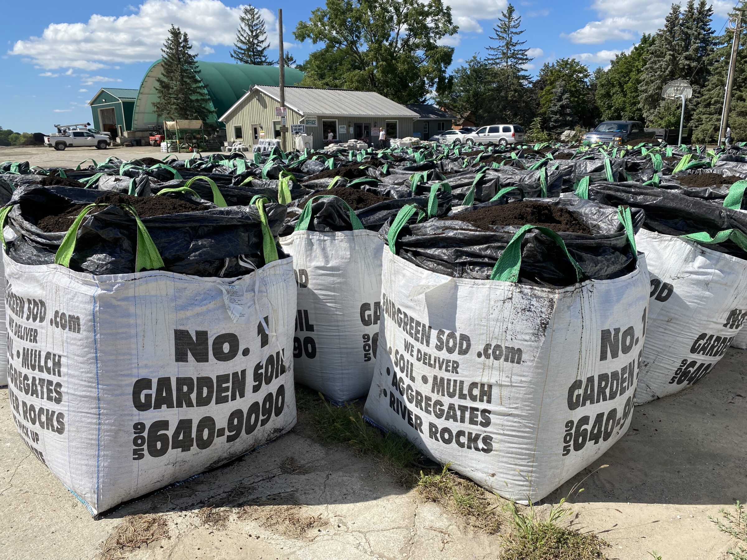 Large bags of garden soil on a concrete lot, surrounded by trees and buildings. Trucks are parked near a green-roofed structure.