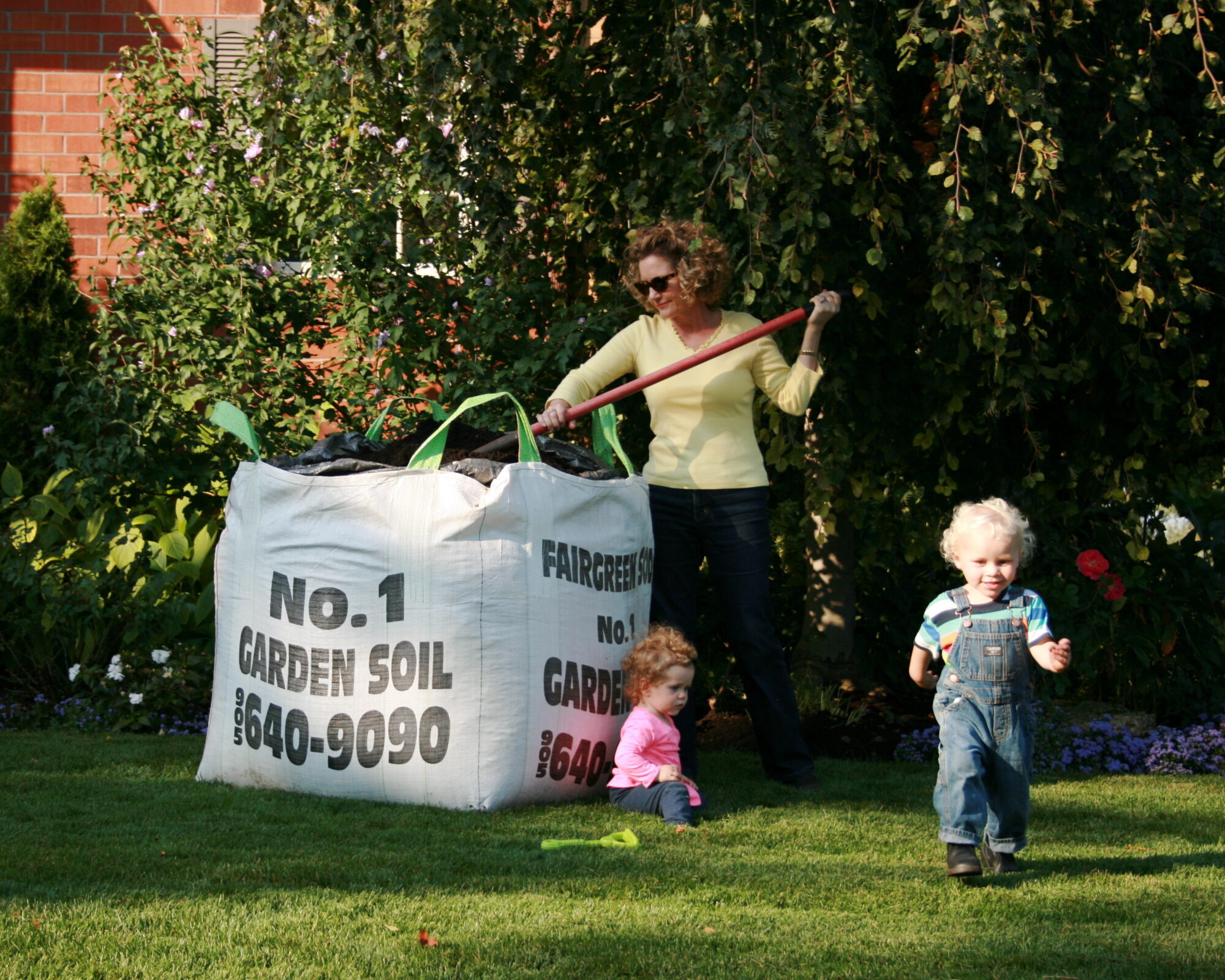 A person tends to garden soil while two children play on the grass, surrounded by green foliage and flowers in a backyard setting.