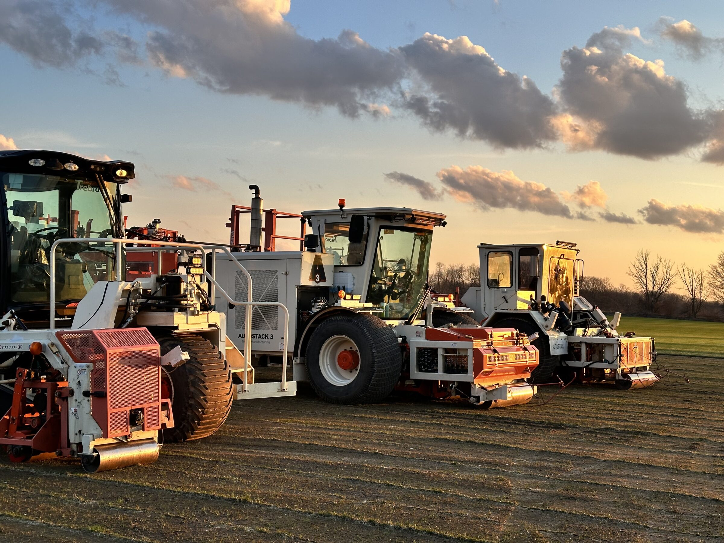 A row of agricultural machinery sits on a field at sunset, with clouds and trees in the background, suggesting a rural setting.