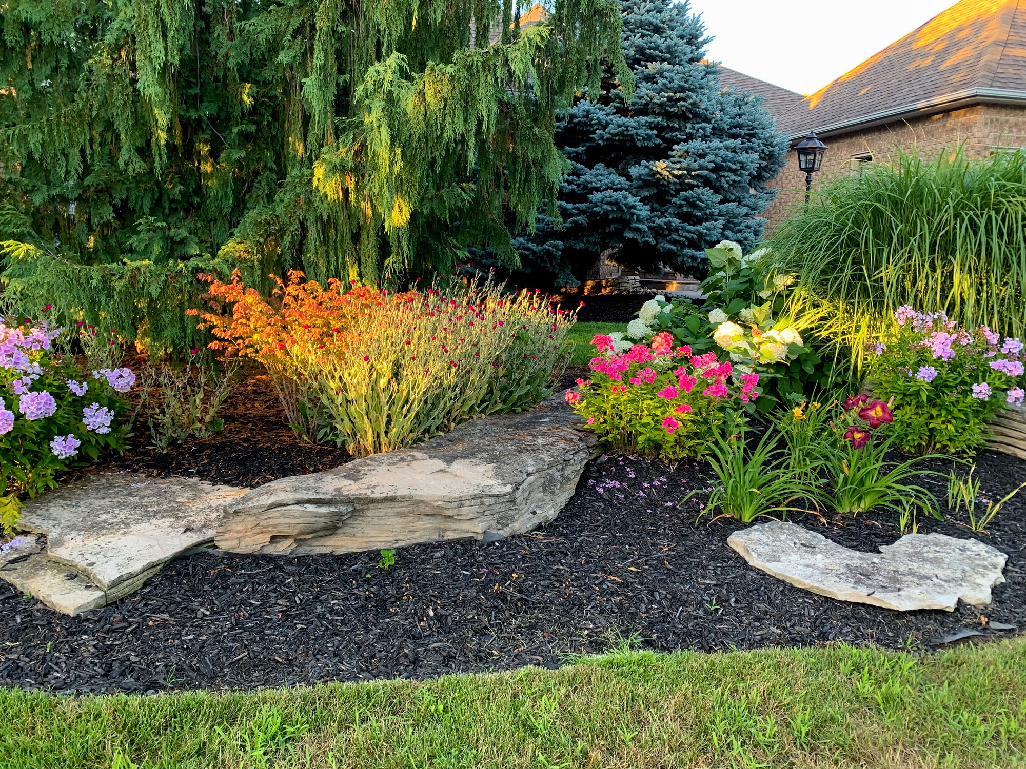 A vibrant garden with colorful flowers, lush greenery, and large rocks illuminated by sunlight next to a brick building with a lantern.