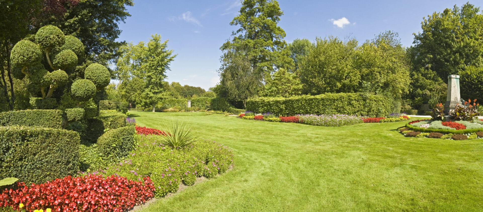 A beautifully landscaped garden featuring topiary, colorful flower beds, and lush green lawns under a clear blue sky, surrounded by dense trees.