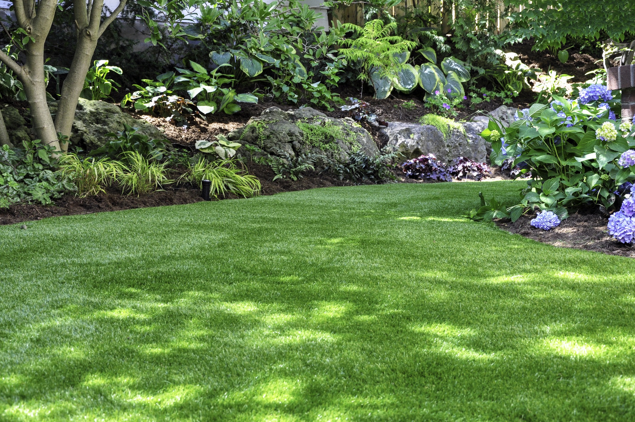 A lush garden with vibrant green grass, colorful flowers, and dense foliage under dappled sunlight, creating a serene and inviting atmosphere.