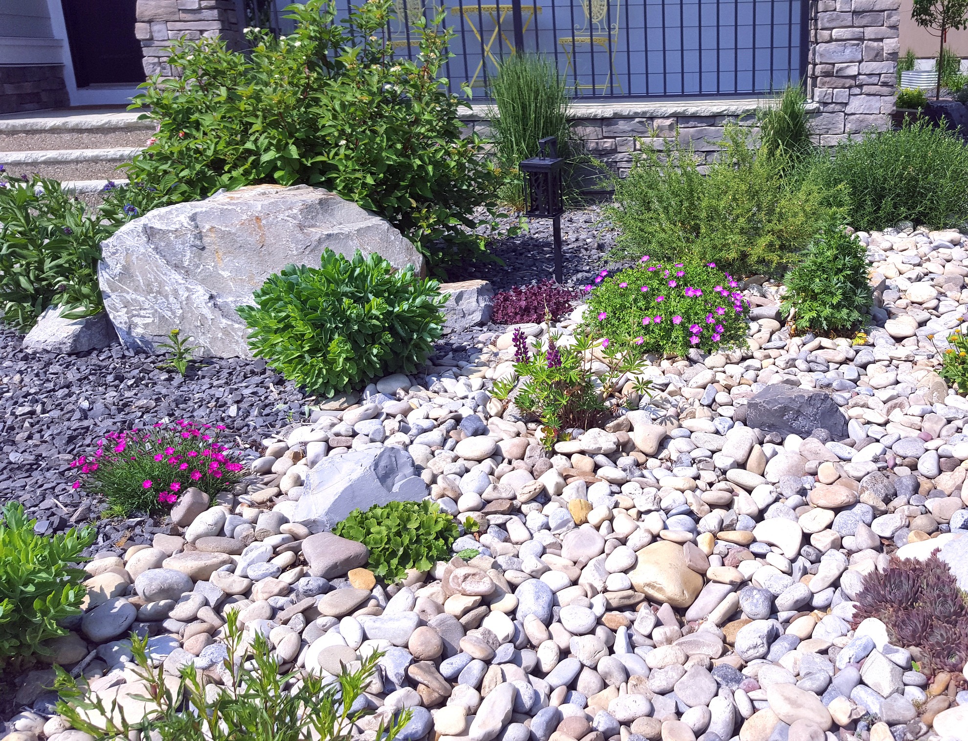 A well-maintained rock garden with various green plants and purple flowers, bordered by a stone wall and a small light fixture.