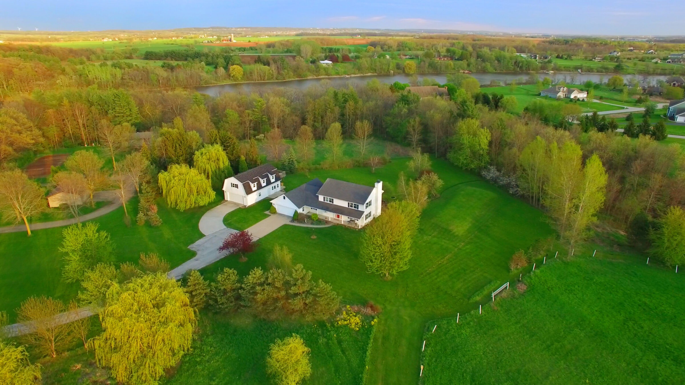 Aerial view of a rural landscape with a white house surrounded by vast greenery, trees, and distant farms near a tranquil river.